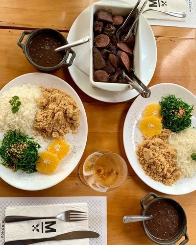 Feijoada servida com arroz, farofa, couve, laranja e a travessa de carnes