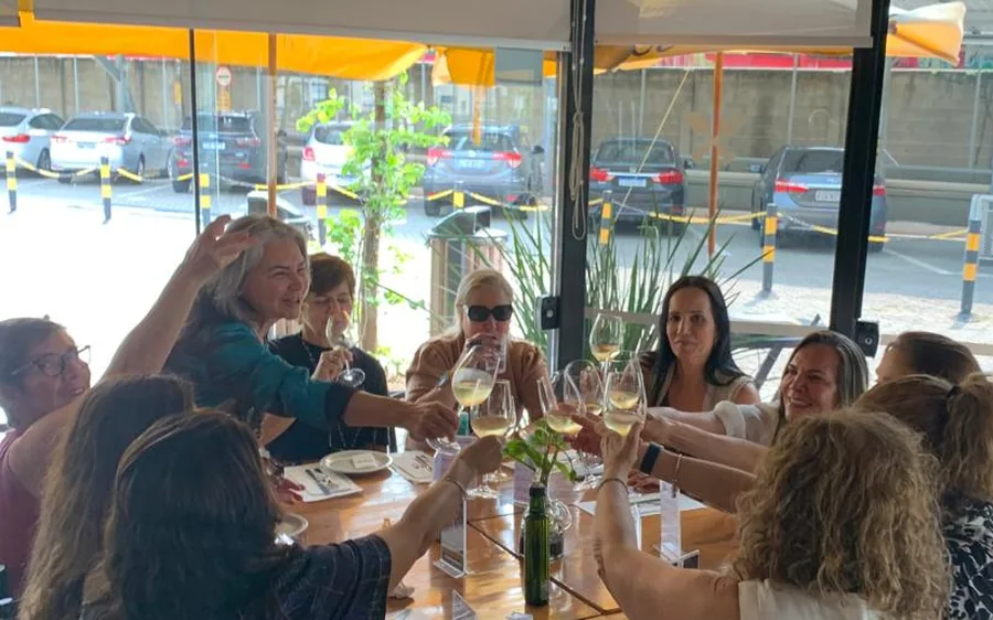 Grupo de amigas brindando com taças de vinho branco na mesa grande do Mironga